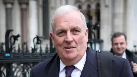 Peter Macdiarmid/Getty Images Kelvin MacKenzie, who has white hair and wears a white shirt, tie, and navy suit, walks on a street with the background blurred out. 