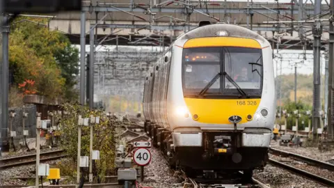 Network Rail A yellow and silver train running on a railway track on a grey overcast autumnal day. Two members of staff sit in the front of the cabin, one is driving the train.
