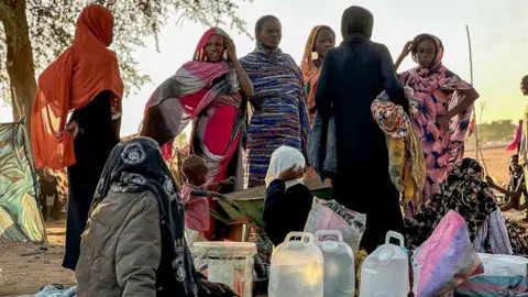AFP via Getty Images Displaced Sudanese who fled el-Fasher after the city fell to the Rapid Support Forces (RSF), rest near the the town of Tawila. Women in colourful scarves are standing up looking like they're wondering what to do next. Some people are sitting on the floor with their belongings.