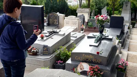 Getty Images A tourist in a windbreaker takes a photo on a digital camera of Edith Piaf's tombstone at the Pere Lachaise cemetery