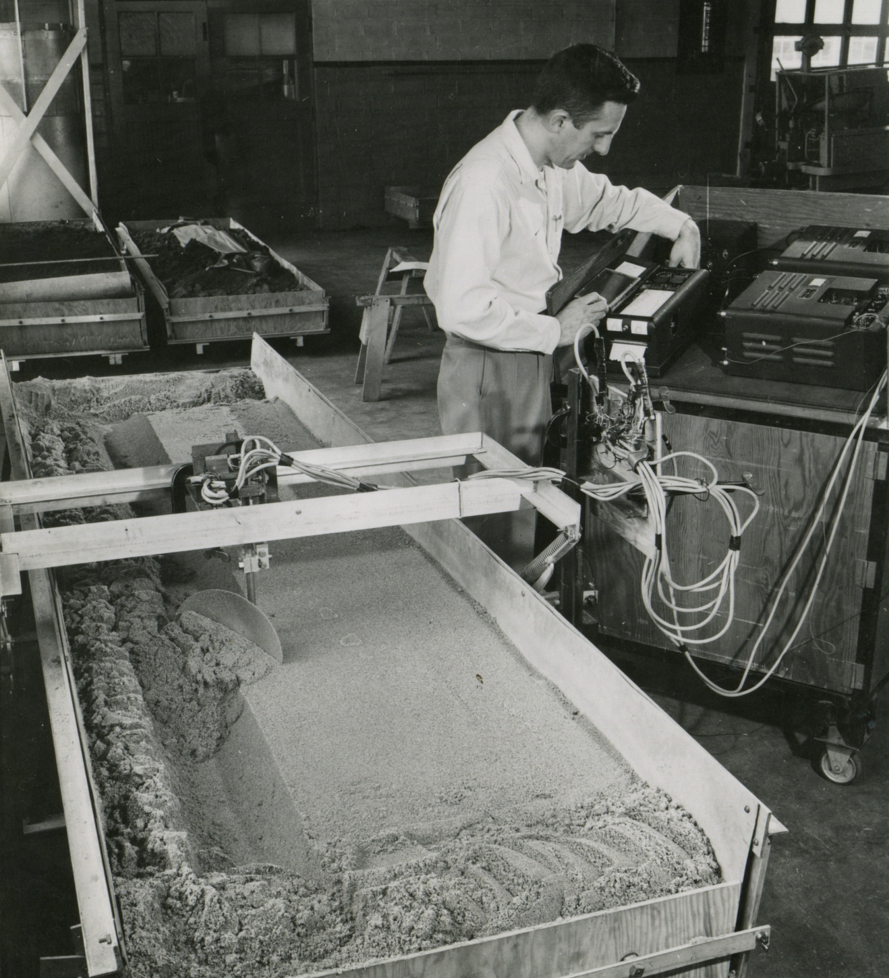 Historical photo of man in white shirt working on controls for machine in laboratory with soil bins