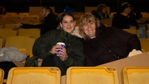 Wolves Foundation Two women sit in yellow chairs in a stadium stand, wearing dark waterproof jackets. They are huddled together and smiling, and the one on the left is holding a coffee cup. The woman on the left has dark hair tied back and the one on the right is blonde. It is dark but the stand is lit.
