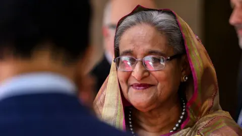 Getty Images Headshot of Sheikh Hasina smiling at an event in Italy in summer 2025. She has grey hair that is largely covered by a colourful headscarf, and is wearing glasses, jewelled earrings and a silver beaded necklace. 