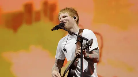 BBC A red-haired Ed Sheeran sings into a microphone while playing a guitar. He is wearing a white tee-shirt