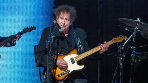 Getty Images Bob Dylan playing a sunburst guitar while dressed in black and sitting on a stool.