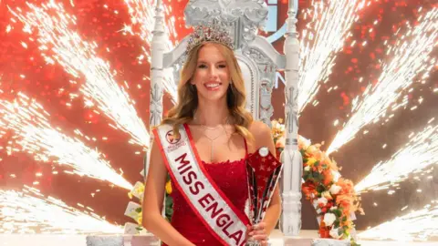 Alan Strutt/Miss England Grace Richardson wearing a red dress and a Miss England sash wearing a tiara and holding a silver trophy