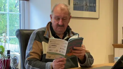 BBC An older man with grey hair is reading a book called Stoke Climsland Parish Retrofit Guide. He is sitting a desk with a laptop in front of him.