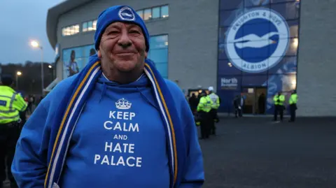 Getty Images A man in a Brighton and Hove Albion beanie and hoodie that says "Keep calm and hate Palace". He is standing in front of a Brighton badge on the outside of the Amex.