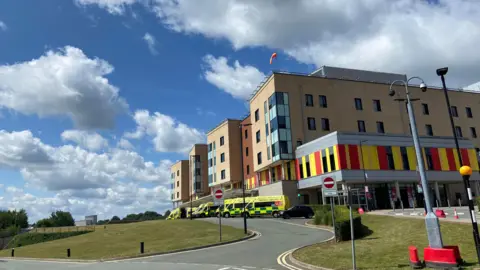 LDRS The outside of the Royal Stoke University Hospital, a five-storey building with red and yellow cladding. There are ambulances parked outside the building. There is a road leading up to the ambulance bays.
