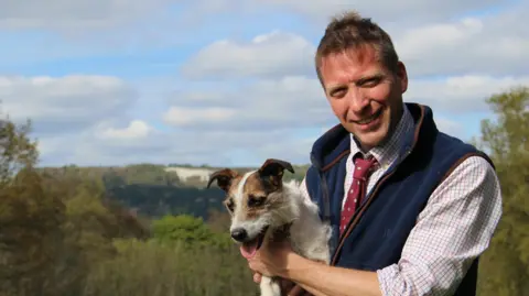 DAISYBECK STUDIOS Yorkshire Vet Julian Norton - who is supporting elected mayor David Skaith's mens' mental health campaign - is smiling at the camera, holding Jack Russell dog Emmy and wearing a blue fleece waiscoat.