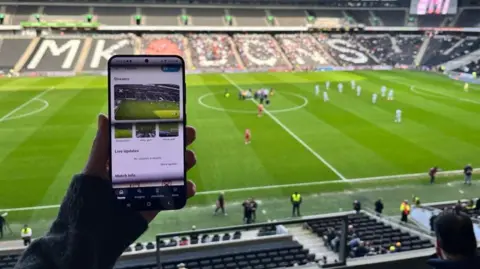 Weaver Labs. Image of someone sitting in a football stadium holding up a mobile phone, with images of a football pitch on it.  In the background can be seen a number of empty stadium seats, a football pitch, football players and officials.