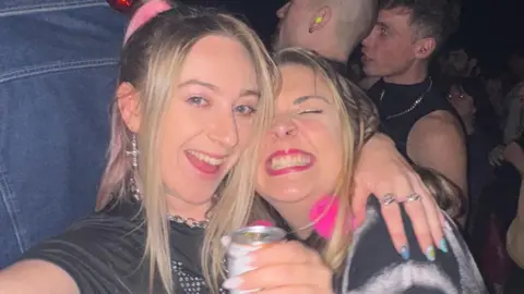 Abi White Abi takes a selfie using a digital camera. She smiles widely at the camera. She has long blonde hair and wears a black crop top with a blue jumper tied round her waist. She has her arm around another woman who has long blonde hair and smiles widely at the camera. She wears a black T-shirt and holds a can of drink.