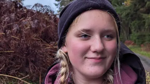 Tara Amelie wearing a dark coloured beanie hat with blonde hair in braids. she is wearing a rain mac over a mauve hoodie and is standing outside on a path. 