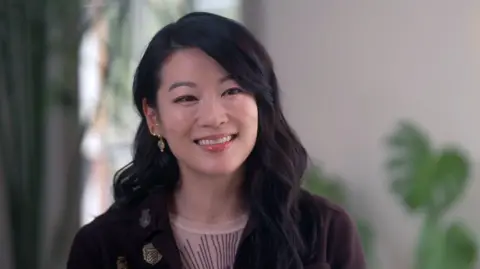 BBC Headshot of Arden Cho smiling to the side of the camera. She has long, wavy, dark brown hair and is wearing several gold, jewelled earrings. She is wearing a sheer cream top with brown embroidery and a brown jacket with gold brooches. She is pictured in a room with cream walls and window behind her, with large cheese plant.