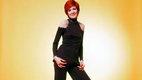 CB Prime LTD A young Cilla Black poses in front of a yellow background. She wears a black jumpsuit with cut out shoulders and a high neck. She has short red hair.