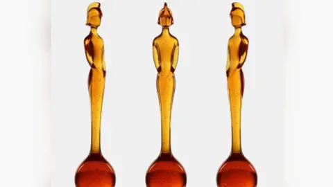 BRIT Awards Three identical statues are displayed side by side against a white background. Each statue is made of translucent amber-colored material with small dark speckles throughout. The statues have an elongated, elegant form resembling a stylized human figure standing upright with arms close to the body. The base of each statue is a rounded, bulb-like shape that gradually narrows into a tall, slender torso and head. The figures have a helmet-like headpiece, giving them a distinctive silhouette.