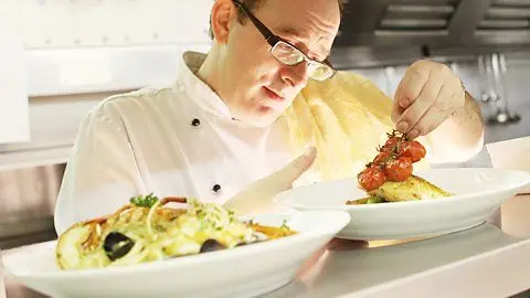 LinkedIn John Quigley in a kitchen. He is dressed in chef's whites, with glasses, and is looking at a plate with food on it in front of him.