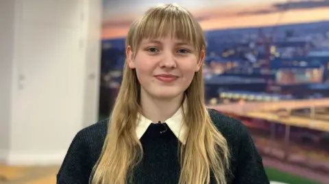BBC A woman with blonde hair smiles into the camera. She is wearing a blue and green coloured jumper with a shirt underneath. She is standing in office in front of an image depicting a city skyline.
