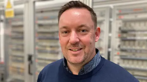 BBC A man smiles at the camera. He has short brown hair and stubble. He is wearing a dark blue collared shirt and dark blue jumper. He is standing in front of glass fronted metal cabinets which have batteries on charge inside.