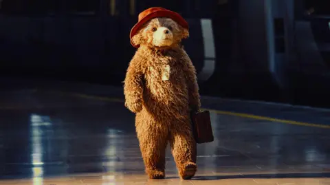 Isha Shah A first look at Paddington in the new West End musical. He's wearing his famous red hat, carrying a suitcase and has a note tied around his neck with string. He is dramatically lit as he walks down a railway platform, his brown and gold fur ruffled. He moves freely, without a human directly operating him or puppet strings.
