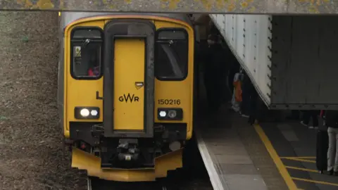 A train on the platform at a railway station. There are people getting on and off of the train from the platform. The train is yellow and black and says 'GWR' on the front.