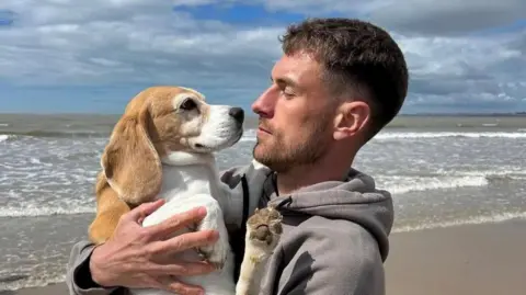 Aaron Ramsey Aaron Ramsey pictured on the beach with sea in background in grey hoodie holding the dog in his arms and looking at it, noses almost touching