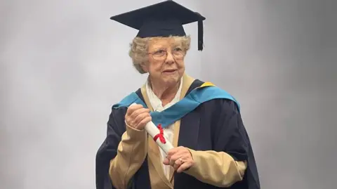 Andrew Gabb An elderly woman with curly grey hair holds a white scroll with both hands while wearing a navy blue gown and navy blue mortarboard on her head. 