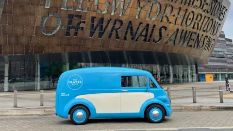 Morris Commercial A retro looking blue and cream van with blue wheel trims is parked outside the Wales Millennium Centre. A logo on the side of the vehicles says 'The Travel Show'.