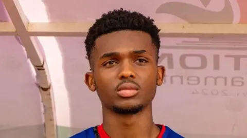 Supplied The head and shoulders of Takondwa, who is wearing a black patterned football shirt with a blue vest over the top. He is facing just beyond the camera lens and appears to have his hand on his chest.