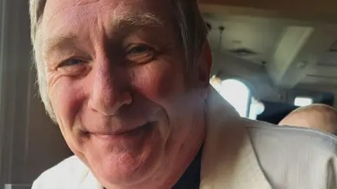 Family A close-up of a man in his 50s. He has a slightly red face and is smiling with his mouth closed. He has sideburns and a white collared shirt-jacket.
