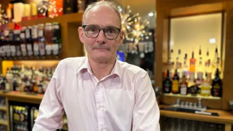 Mark Milne is a balding man with black-rimmed plastic spectacles and a pink open-necked shirt. He is standing in front of a lit bar gantry full of bottles and some Christmas decorations. He is smiling slightly.