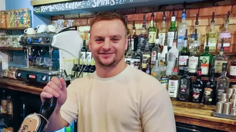 BBC Rob Hardie with brown hair and white t-shirt pulling a pint behind the bar with alcoholic drinks and a coffee machine in the background.