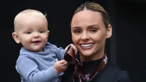 Chloe McNiven A woman (Chloe) is smiling while holding her blonde haired baby boy. The medal is around her neck, and her baby is holding it in his hands while smiling toothlessly. 