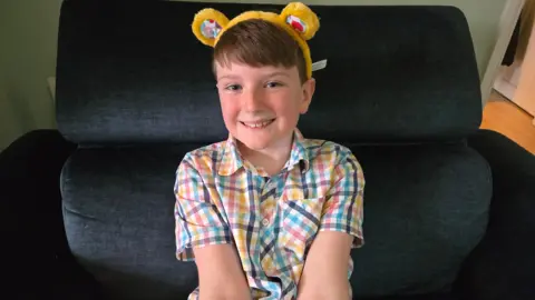 BBC Taylor wearing a gingham, multi-coloured shirt and Pudsey ears smiling at the camera while sitting in a large dark armchair.