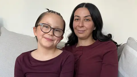 BBC A woman in her 30s with dark long wavy hair, olive skin and brown eyes sits next to a girl of 10 with short brown hair which is braided on top, and she is wearing black framed glasses. They are sitting on a grey sofa both smiling at the camera  and are both dressed in burgandy coloured tops, the wall behind them is painted cream. 