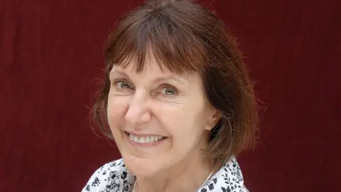 Getty Images A headshot of a middle-aged woman smiling at the camera.  She has dark hair and brown eyes,  She is wearing a white blouse with a black flower print on it.
