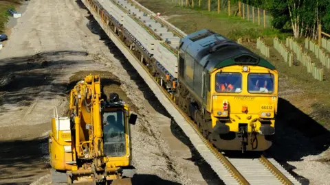 East West Rail A yellow trail carriage on a railway line, that is being built, next to another piece of machinery, that is yellow in colour. There are trees to the left, that have been planted and rubble on the ground. 