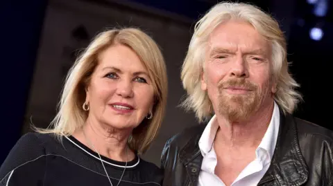 Getty Images Joan Templeman and Sir Richard Branson look towards the camera. She has long blonde hair and a dark top. Branson has light long hair, a beard, white shirt and brown leather jacket. 