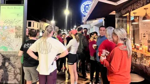BBC A mixture of men and women wearing running gear, stood round socialising. Some have beers in their hands. They're stood outside a bar at night time. There is a sign post showing a map of Bath Road by the side of them. 