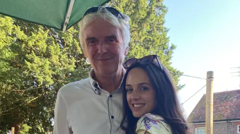 Family handout A man with short white hair wearing a white shirt and sunglasses on his head. He is stood next to his daughter, who has brown hair and is wearing a floral top and sunglasses on her head.