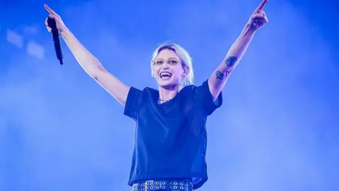 Getty Images Emily Armstrong holding her arms up on stage. She is wearing a black t-shirt and black belt. She has blonde hair tied up, she is wearing yellow glasses. 