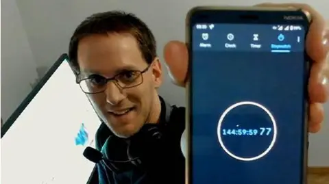 Guinness World Records A man wearing glasses, holding a phone to the screen which reads "144:59:59:77".