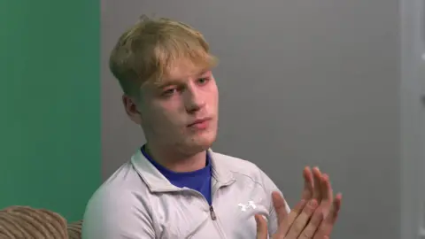 BBC A blonde-haired teenage boy with a grey zip up jumper on, rubbing his hands together whilst looking into the distance.