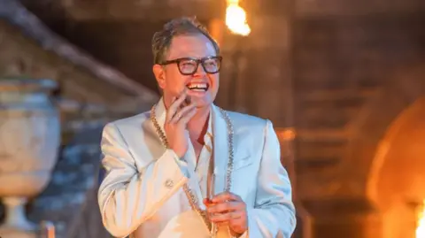 BBC/Studio Lambert/Paul Chappells Alan Carr in a white jacket with a jewelled collar and white shirt. He is holding a glass of champagne and his other hand is up to his face. He is smiling and looking to the side of the camera. It is night-time and behind him blurred out is The Celebrity Traitors' castle, with flamed lanterns lighting up the surroundings. Alan is wearing his trademark large framed black glasses and he has short dark hair.