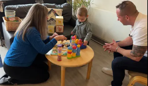 BBC/Tristan Pascoe A child plays with pop up toys with support worker, Teri Hill. The child's father, Chris Bott watches on