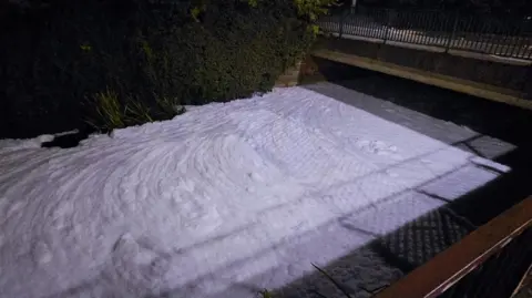 Chris Davison A thick layer of white foam with ripples in it is completely covering the surface of a river. There is a bridge in the background. The photo appears to have been taken in darkness.