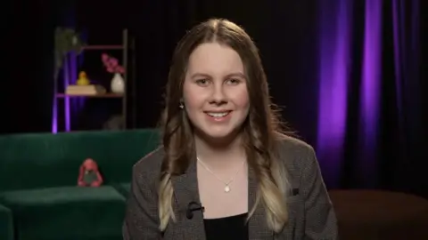 BBC A teenager with long brown hair and beautiful blue eyes smiles into the camera. A little pink cuddly dog sits on the green sofa behind her.