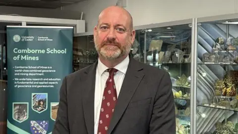 Camborne and Redruth MP Perran Moon stands in front of several cabinets with rocks and minerals inside them. He is bald with a greying beard. He is wearing a grey suit with a white shirt and burgundy tie.