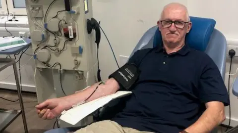 PA Media A man with glasses, a dark shirt and beige shorts, sits on a chair in a doctor's office while donating blood. The machine is standing next to him.