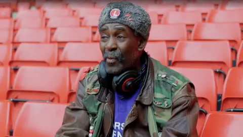 Charlton Athletic Norman Barker wearing headphones, a purple Charlton shirt, a grey hat and camouflage jacket 